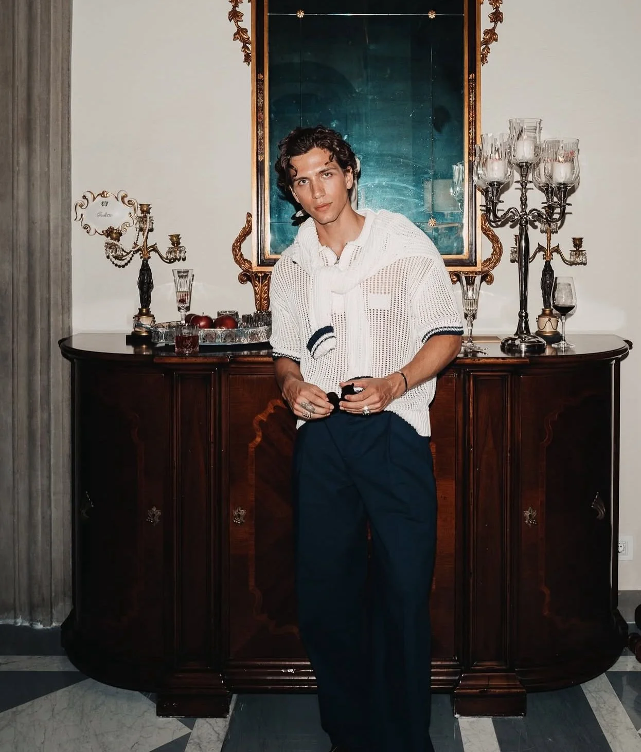 A young man with curly dark hair wearing a white sweater with navy accents, posing in front of an antique sideboard with decorative items, a large mirror, and empty candlesticks behind him.