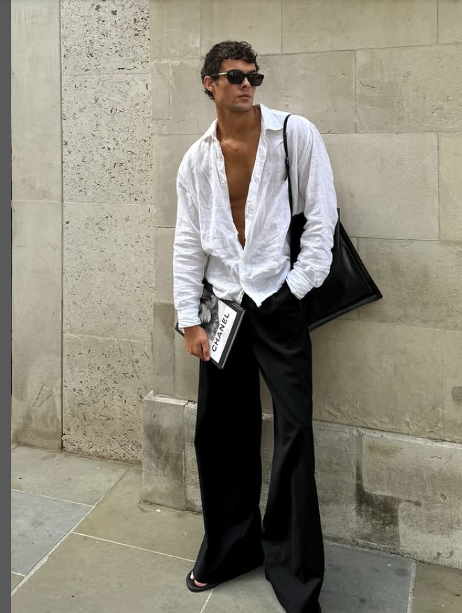 Person wearing a brown leather jacket and baggy jeans, standing in front of a red door, holding a brown bag.