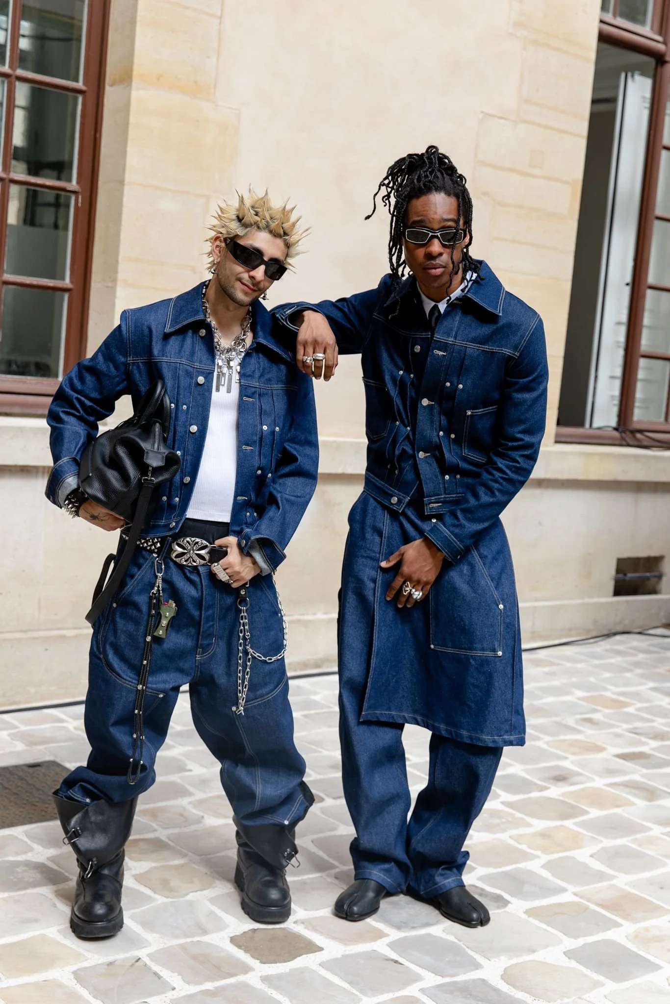 Two young men wearing denim outfits and sunglasses standing on a cobblestone street in front of a beige building with windows.