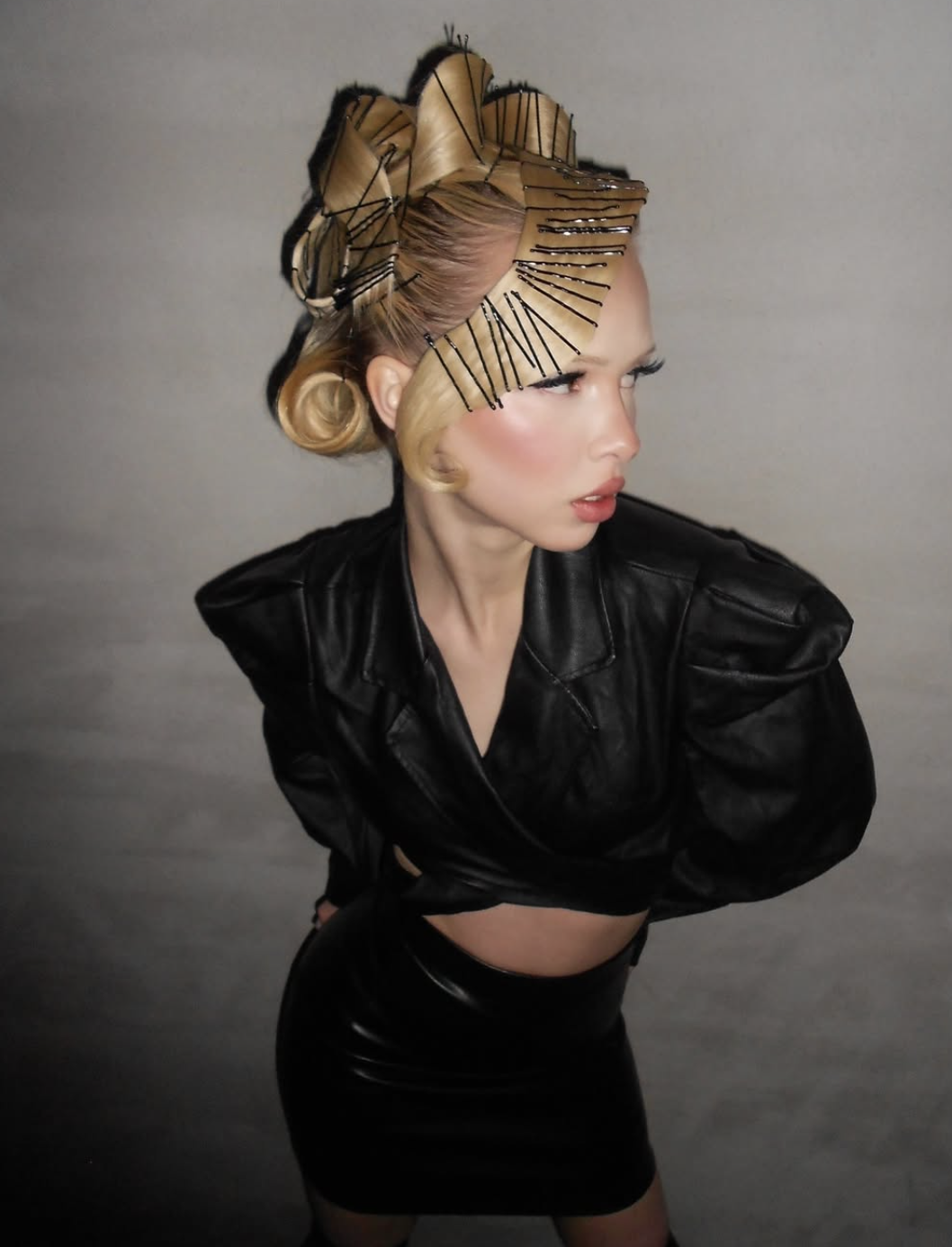 Person with avant-garde hairstyle and makeup wearing a black leather outfit, looking off to the side against a gray background.