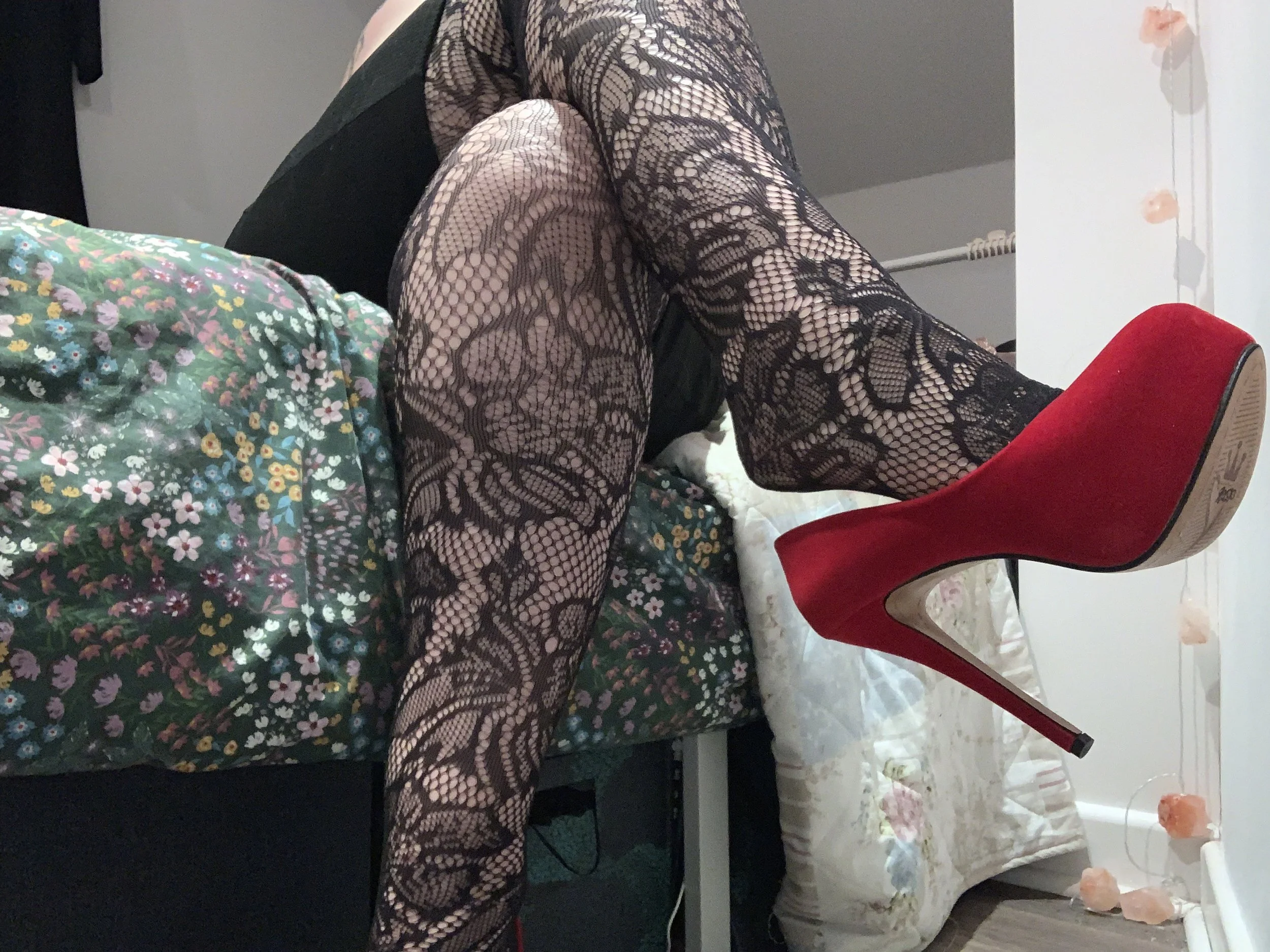 A person wearing black lace sheer tights sitting on a bed with floral bedspread, wearing red high heels. 