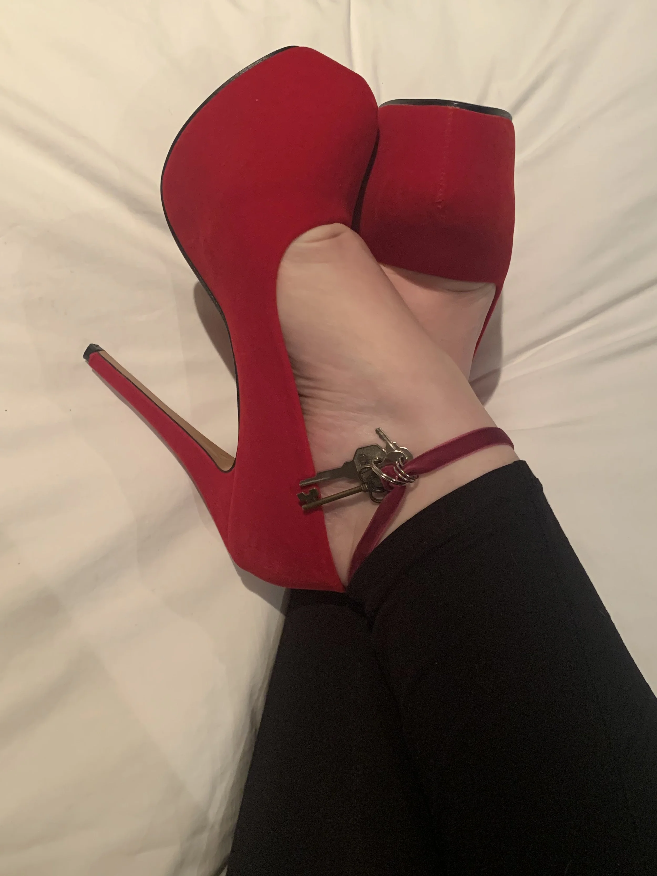 A person wearing black pants and a red high-heeled shoe with a key around their ankle. The person is lying on a white surface.