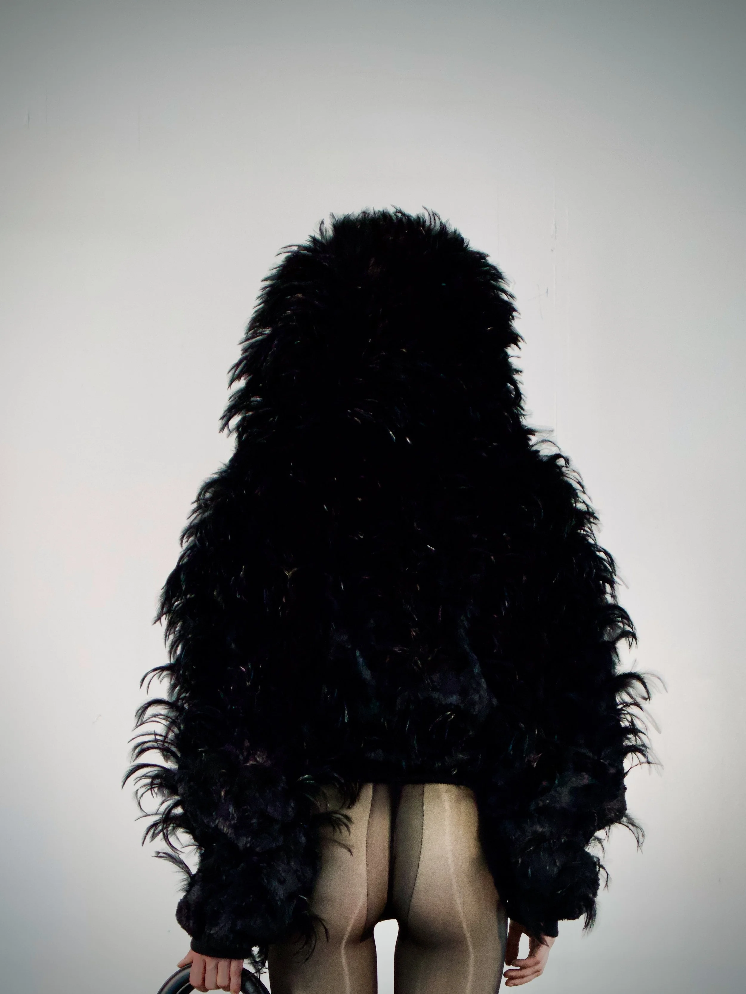 A person standing against a plain white wall, wearing beige pants and a large black fur coat. They are holding a black handbag in one hand.