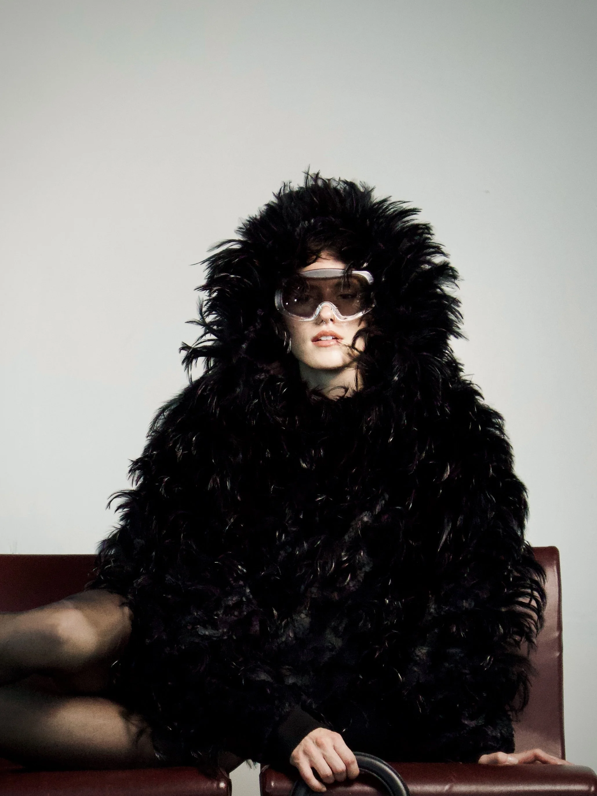 A woman wearing a large, fluffy black coat with a hood and clear safety goggles. She appears to be sitting on a leather bench with her left hand resting on a wheelchair handle, in a room with a plain light-colored wall.