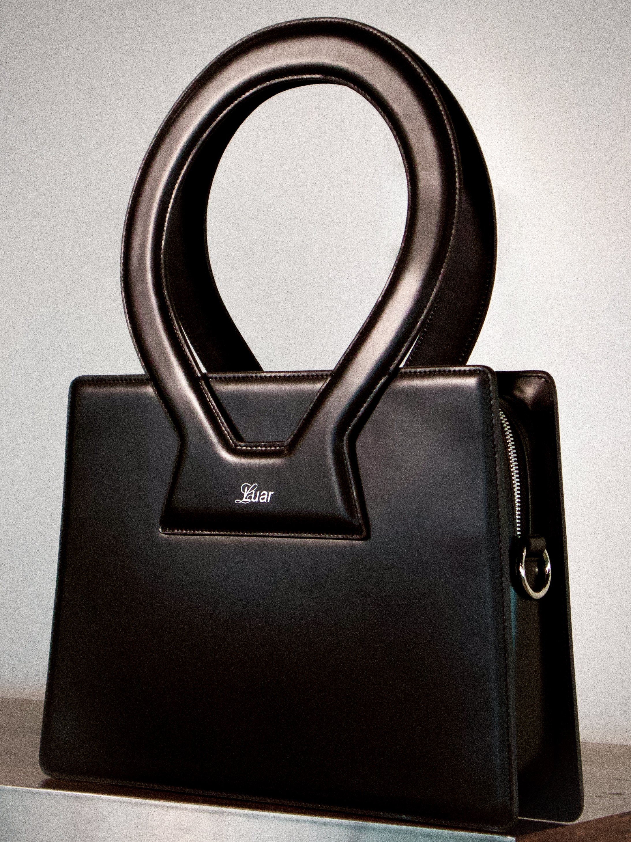 Black leather handbag with the word "Luar" embroidered on a patch on the front, positioned on a gray surface against a plain background.