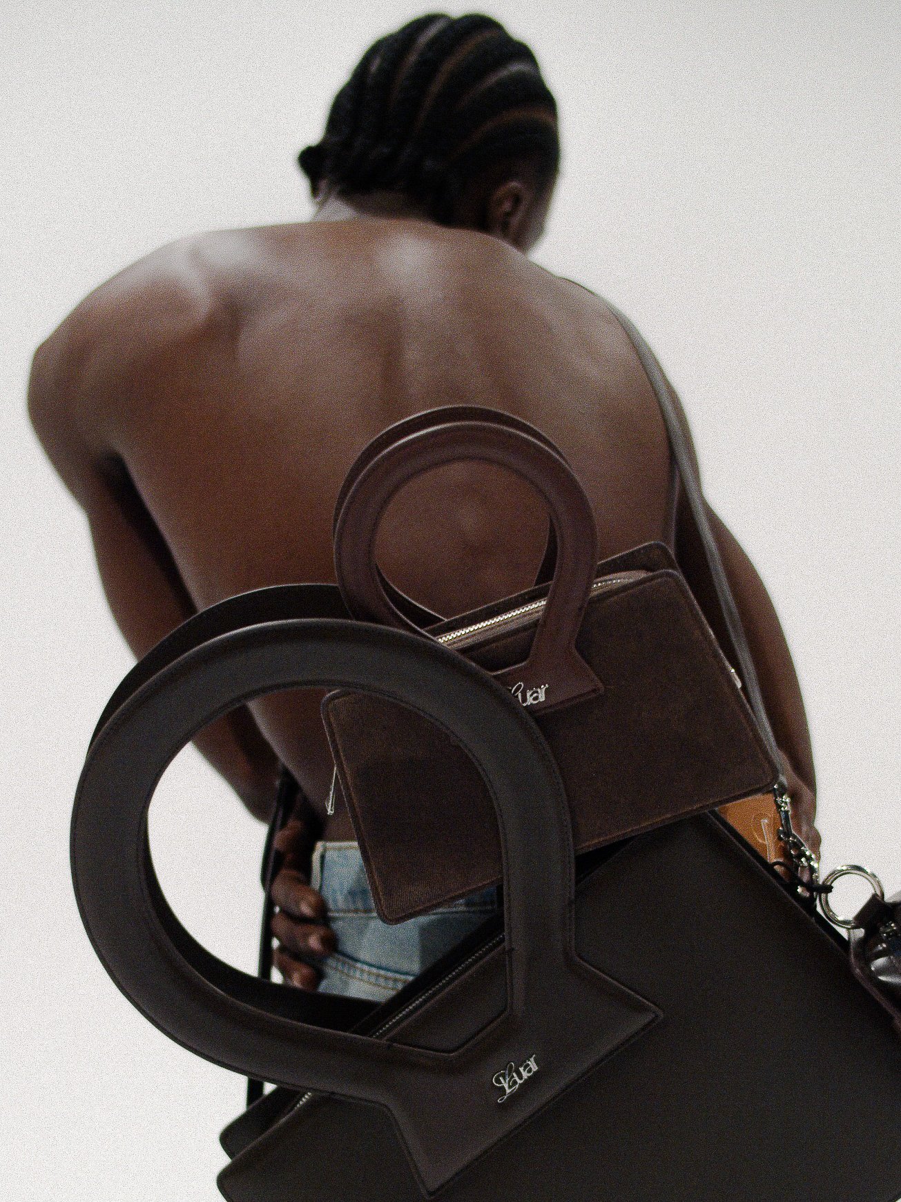 A shirtless person with braided hair, carrying a brown handbag on their shoulder, seen reflected in a circular mirror attached to the handbag, with a white background.