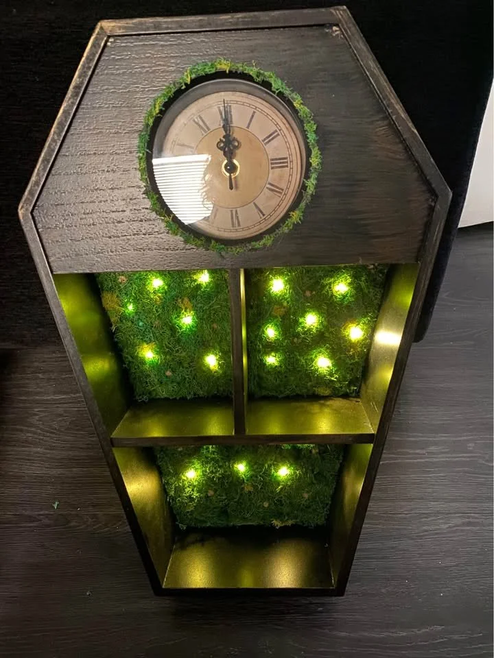 A wooden hexagonal clock with a round face, Roman numerals, and a pair of black clock hands, decorated with a green moss wreath around the face. Below, there are three open compartments with green moss and small yellow-green lights inside.