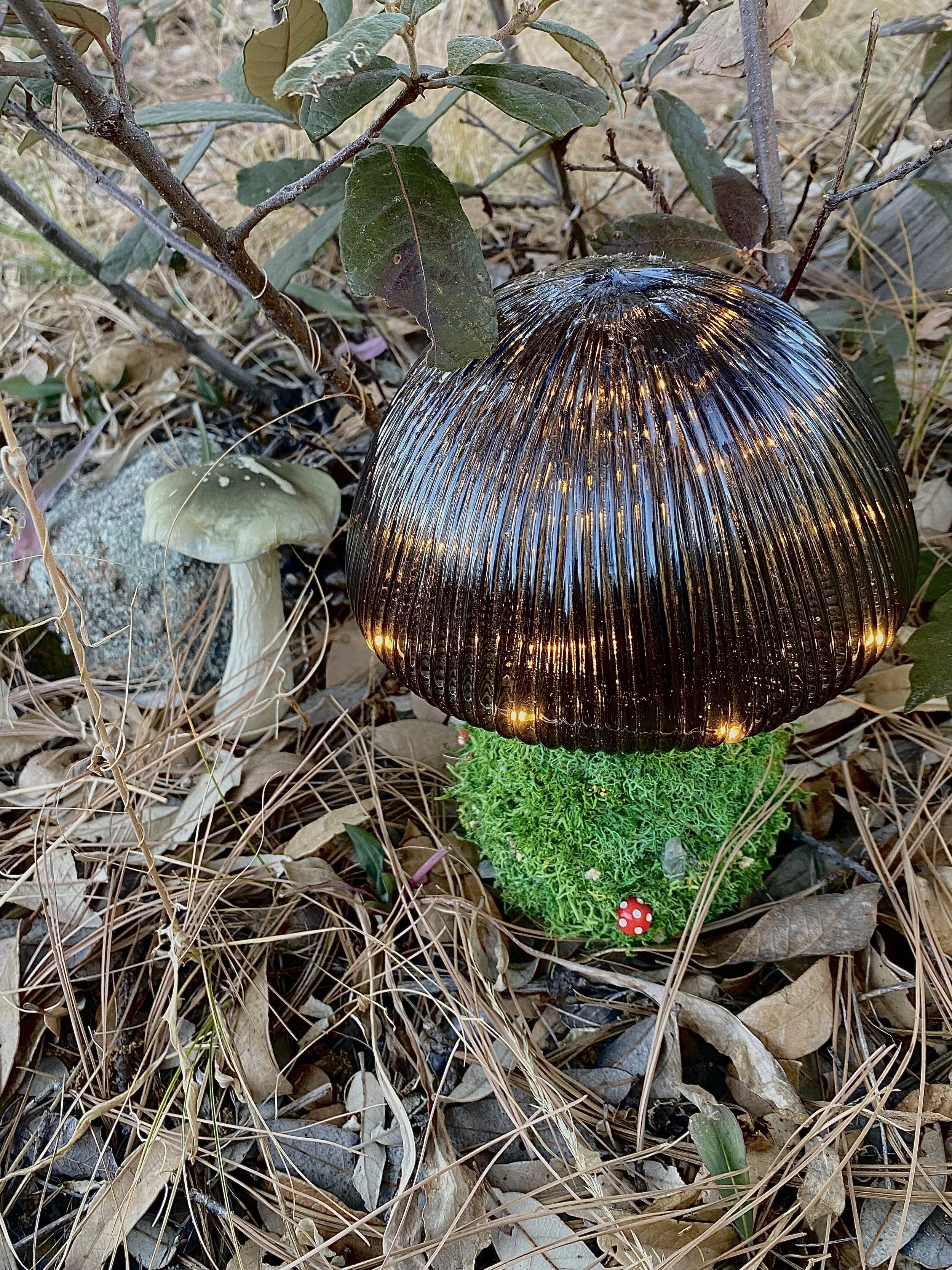 A decorative mushroom sculpture with a glossy, black, ribbed cap and a green moss-like base, surrounded by fallen leaves, a small red and white mushroom figurine, and an earth-toned outdoor setting.