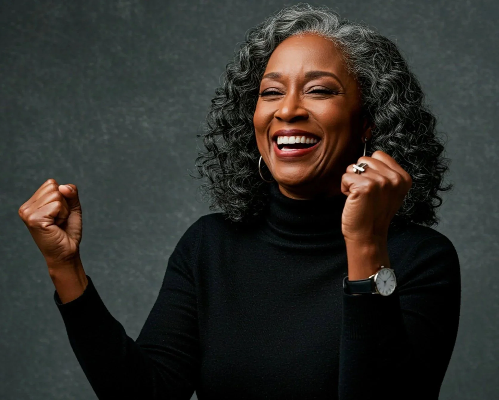 An older woman with curly gray hair smiling, wearing a black turtleneck, a wristwatch, earrings, and a ring, raising her fist in a gesture of victory or empowerment, against a dark gray background.