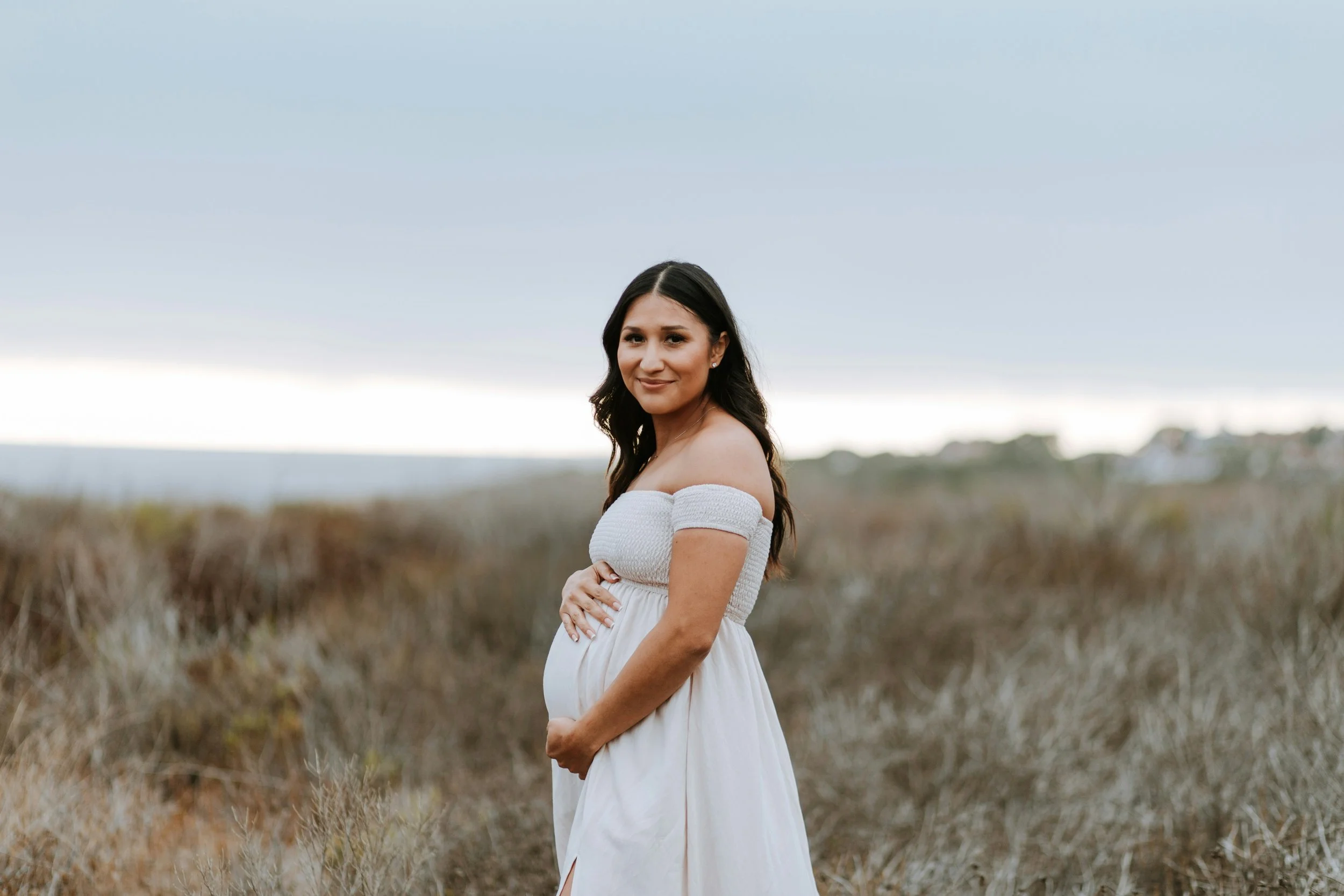 Pregnant woman in a white dress standing in a coastal field, prenatal care by Birthing by Nature in Ventura County, CA