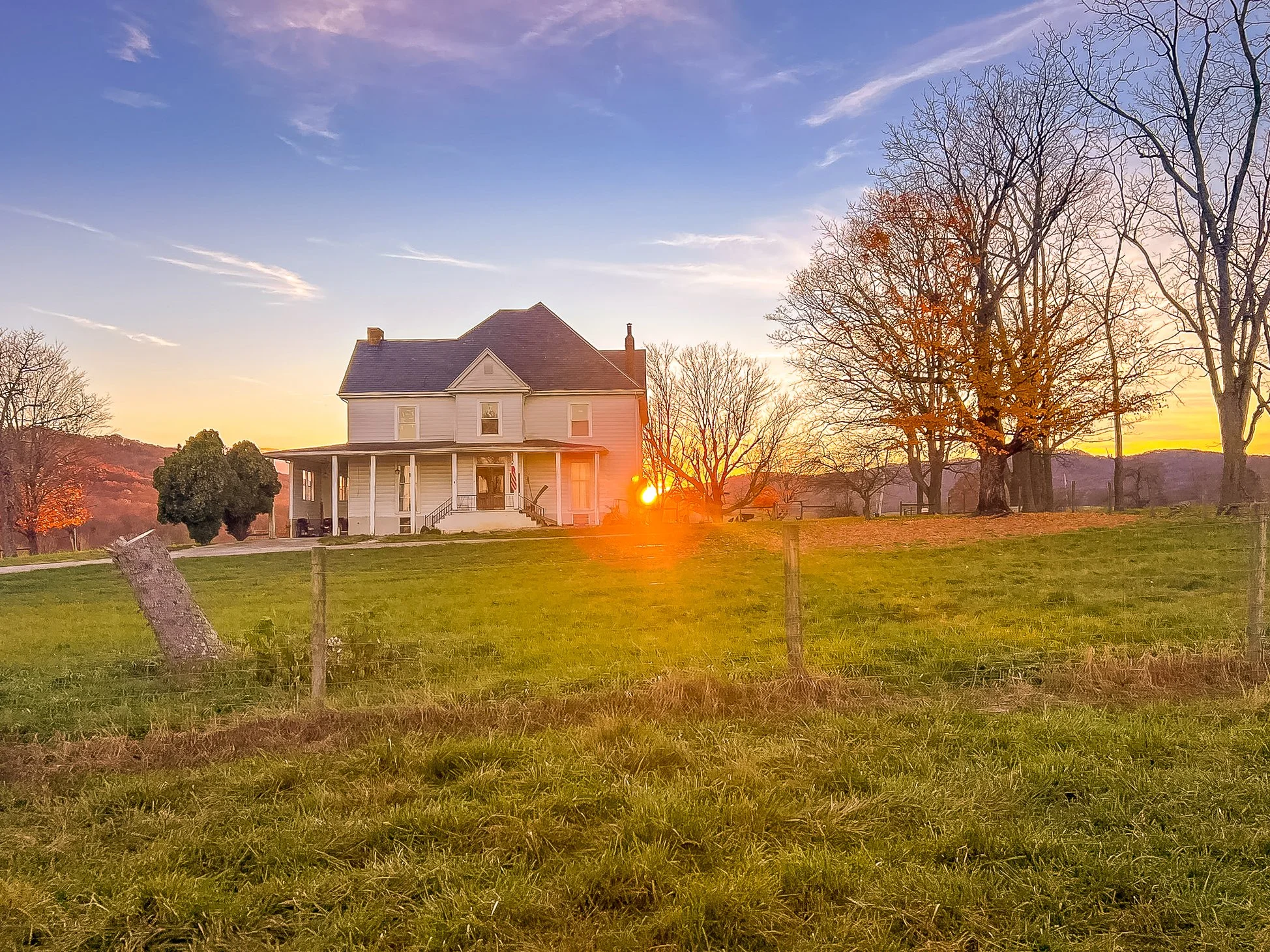 Sunset over a white, two-story house on a farm with green grass and leafless trees.
