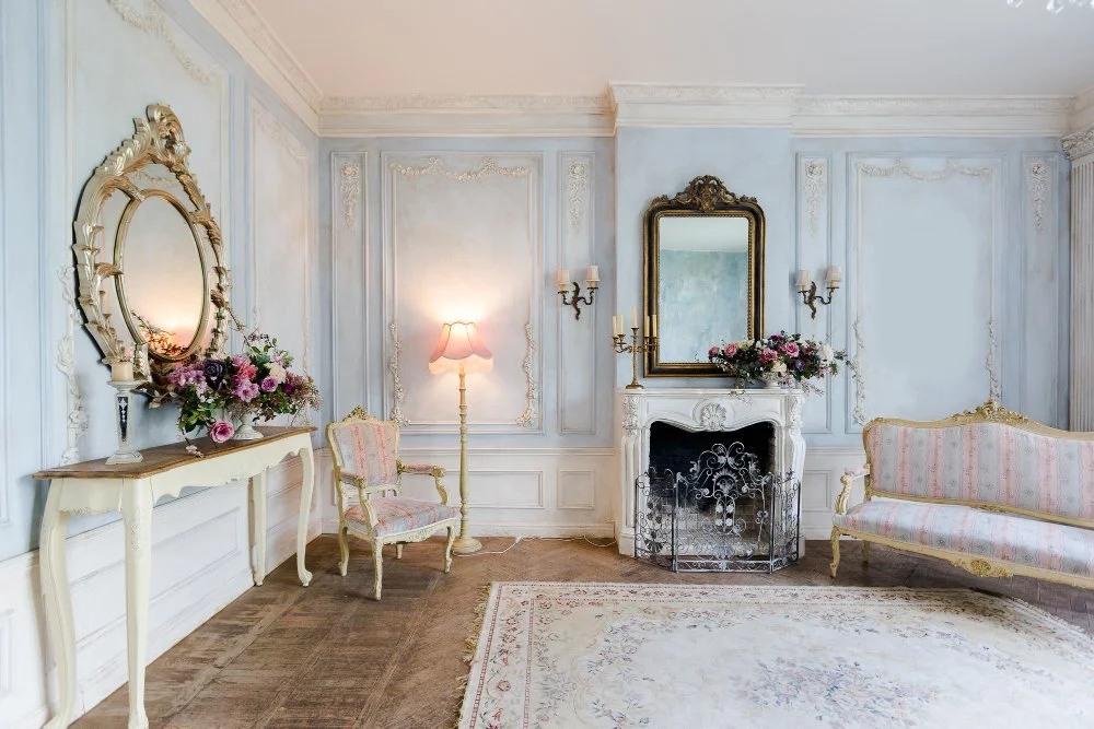 Elegant vintage living room with white and gold furniture, floral arrangements, a mirror, a fireplace, and pastel-colored walls.