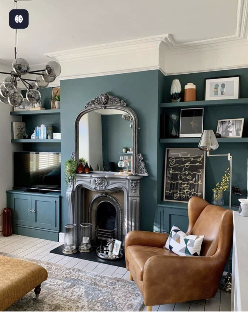 A cozy living room with teal colored walls, a decorative silver mirror above a gray ornate fireplace, a brown leather armchair with a geometric pillow, a mustard-colored ottoman, a black TV on a teal cabinet, and shelves with decorative items, books, and framed artwork.