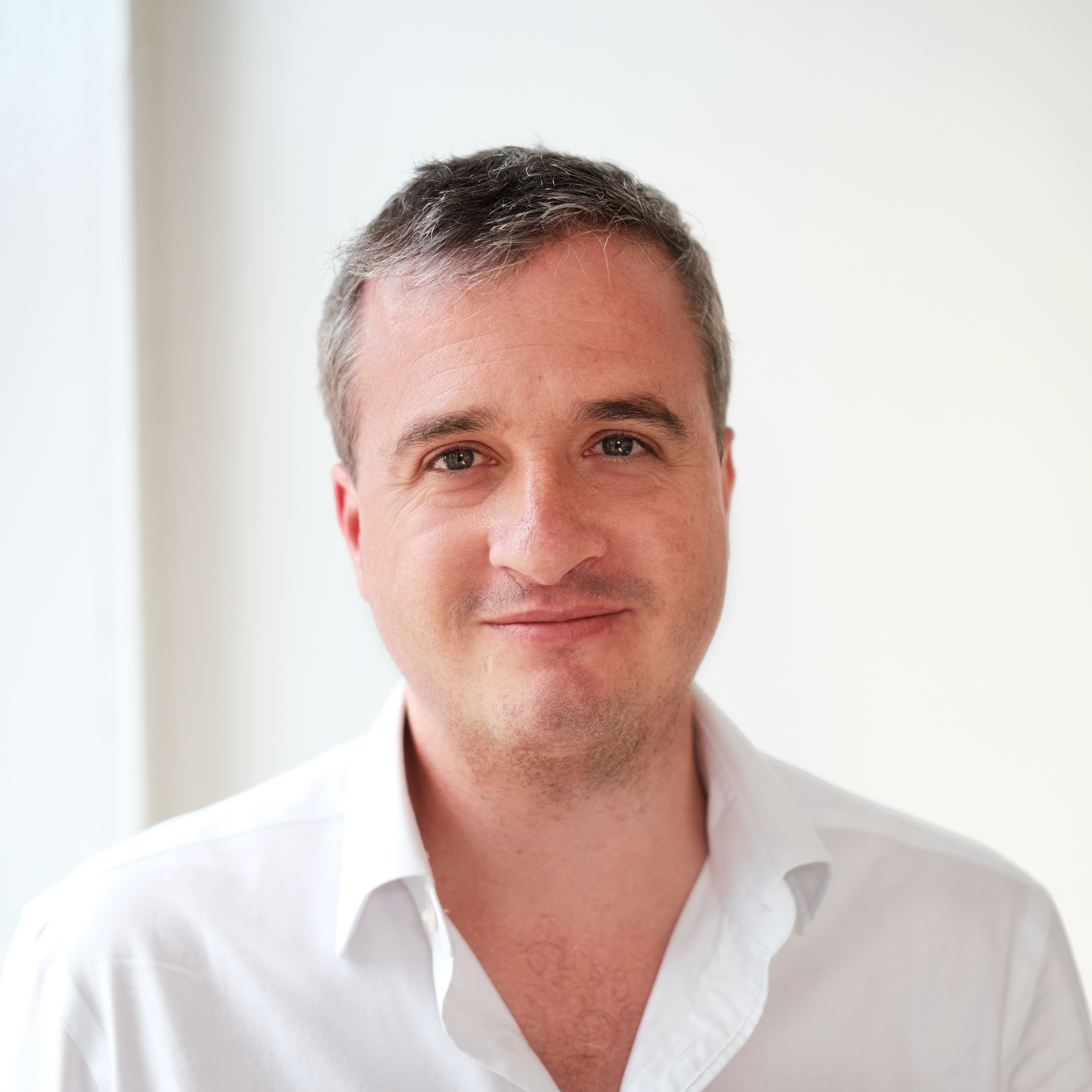 Close-up of a man with short brown hair, wearing a white collared shirt, smiling, against a light background.