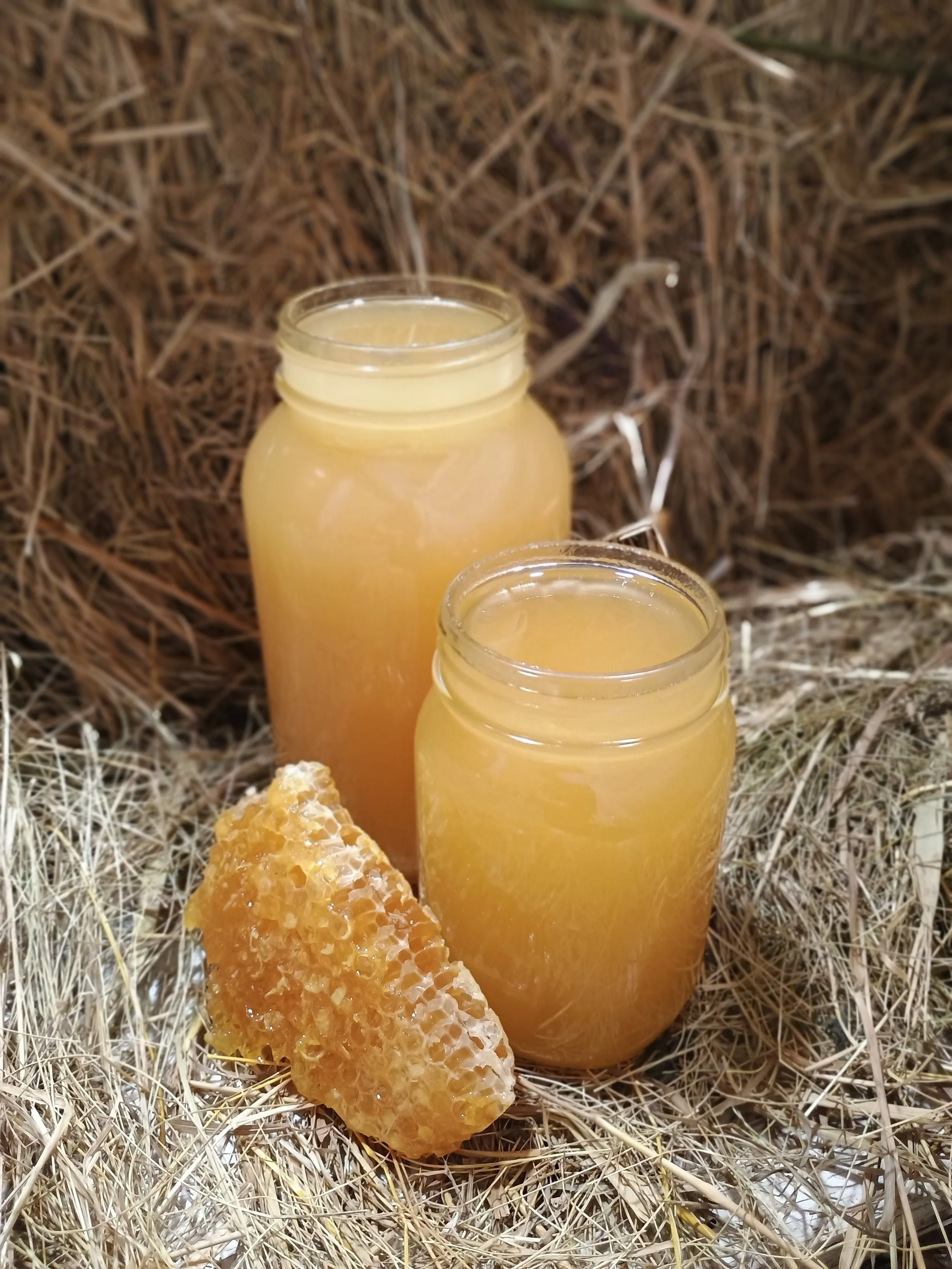 Two jars filled with honey placed on a bed of dry straw, with a honeycomb piece resting in front of them.