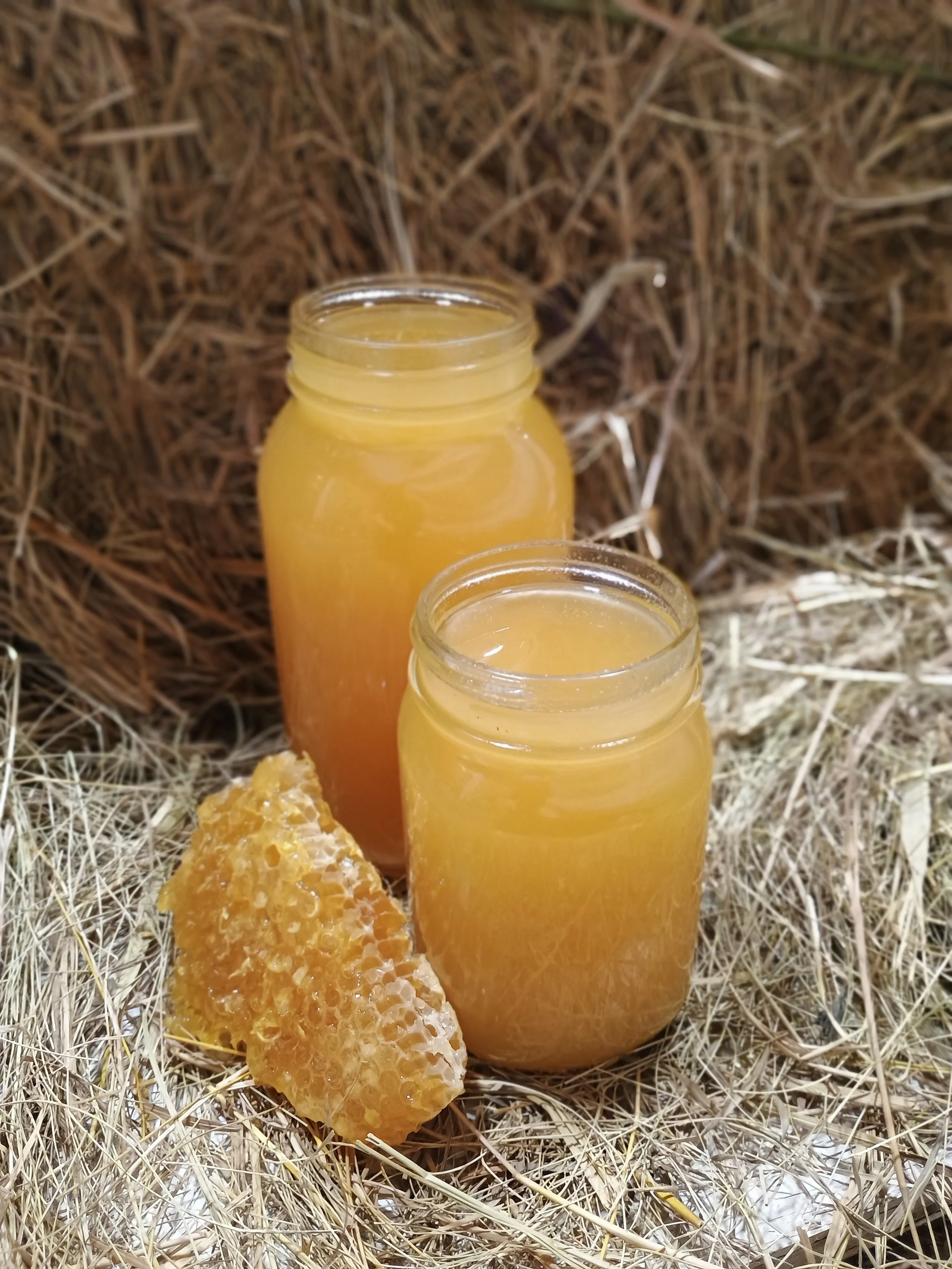Two glass jars filled with honey, one honeycomb piece placed beside them, all set on a bed of dried straw or hay.