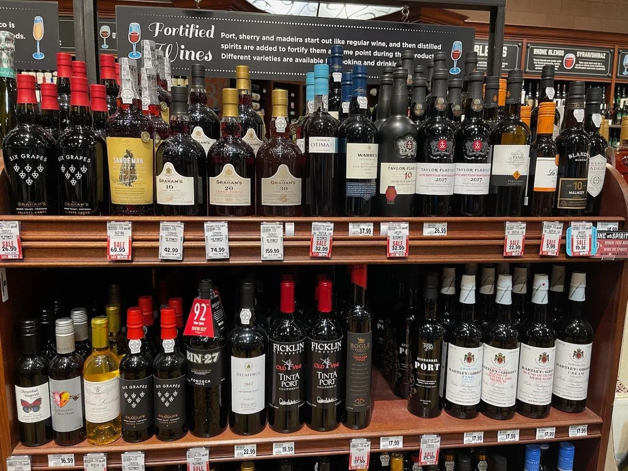 Display of various bottles of red, white, and rosé wines on store shelves with price tags and sale signs.