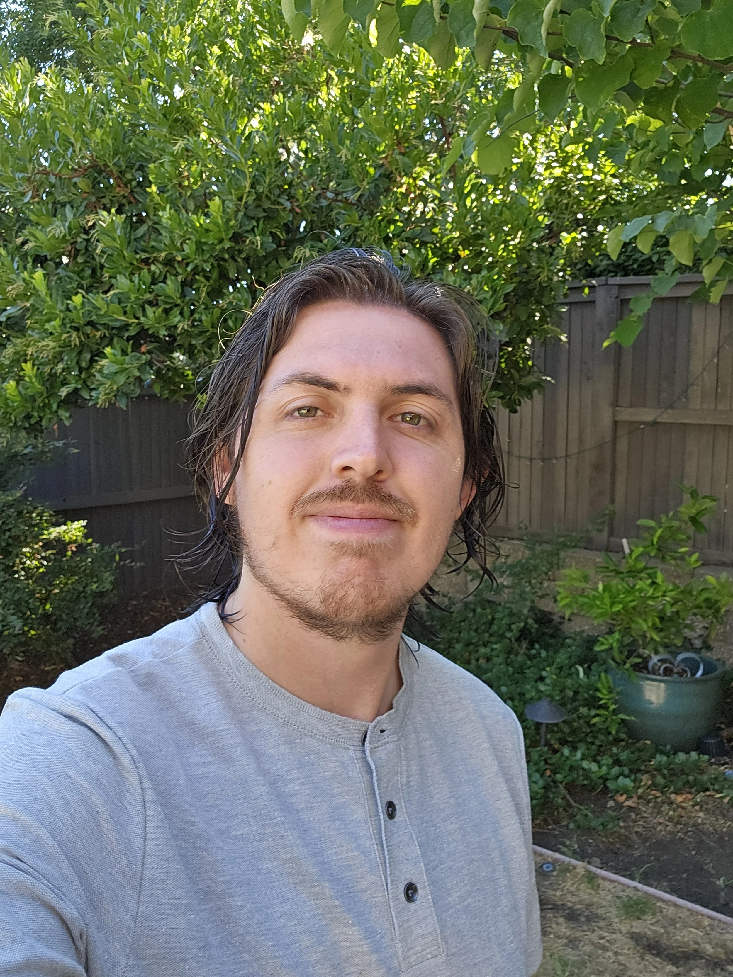 A young man with medium-length dark hair, light facial hair, and a slight smile taking a selfie outdoors in a backyard with green trees, bushes, and a wooden fence.