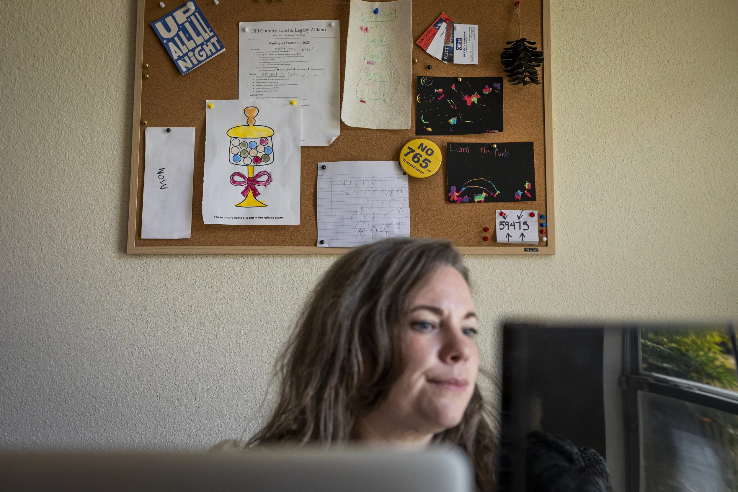 A “No 765 KV transmission” pin is displayed on a bulletin board alongside drawings from Liam Sarot, 9, and Thomas Sarot, 4, while Mia continues to work on responding to emails inside her office in Burnet, TX on Aug. 11, 2026. 