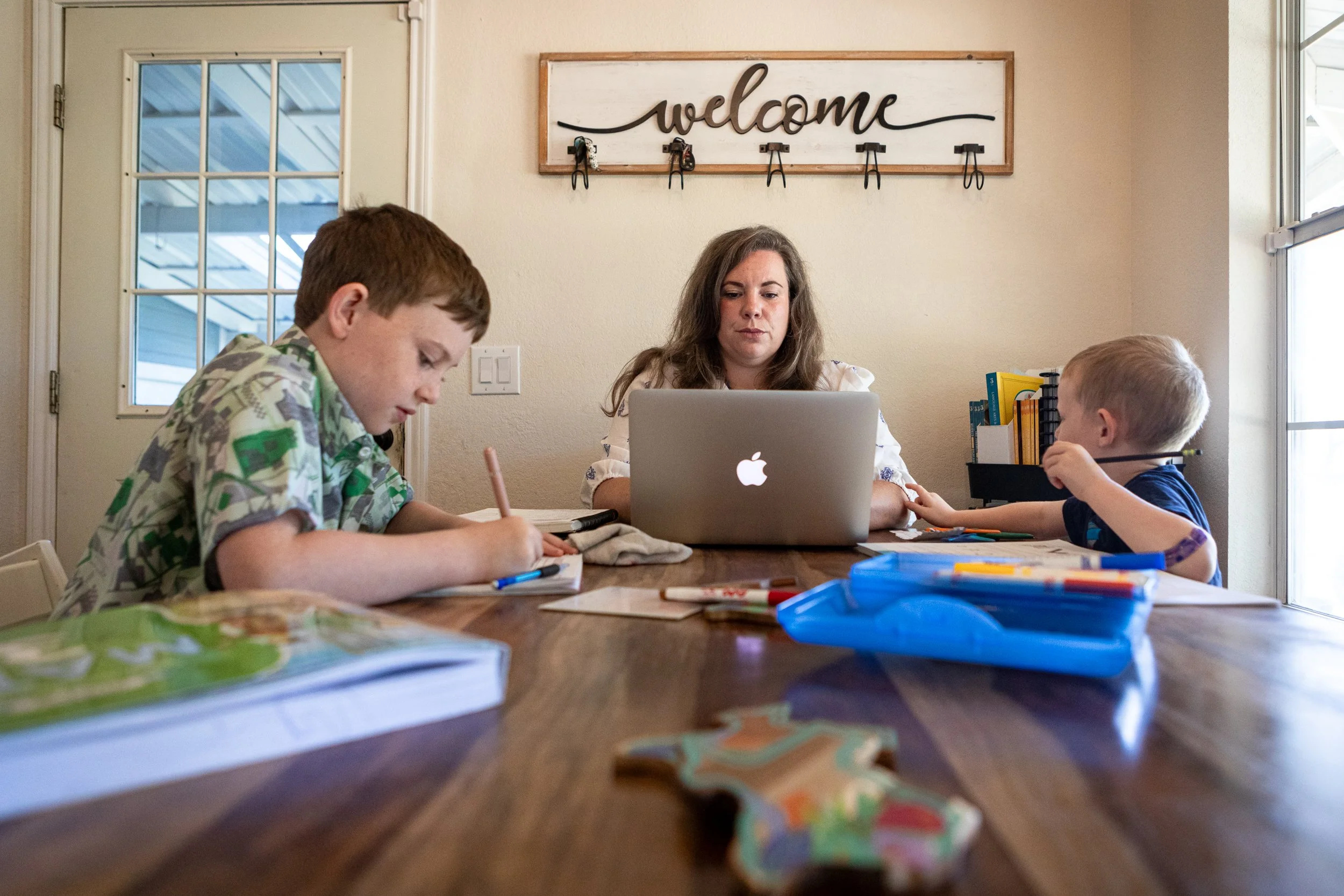 Stay at home mom and lead organizer for the Hill Country Land and Legacy Alliance Mia Sarot drafts and responds to emails while her two kids Liam Sarot, 9, and Thomas Sarot, 4, work on coursework on the kitchen table in Burnet, TX on Aug. 11, 2026.
