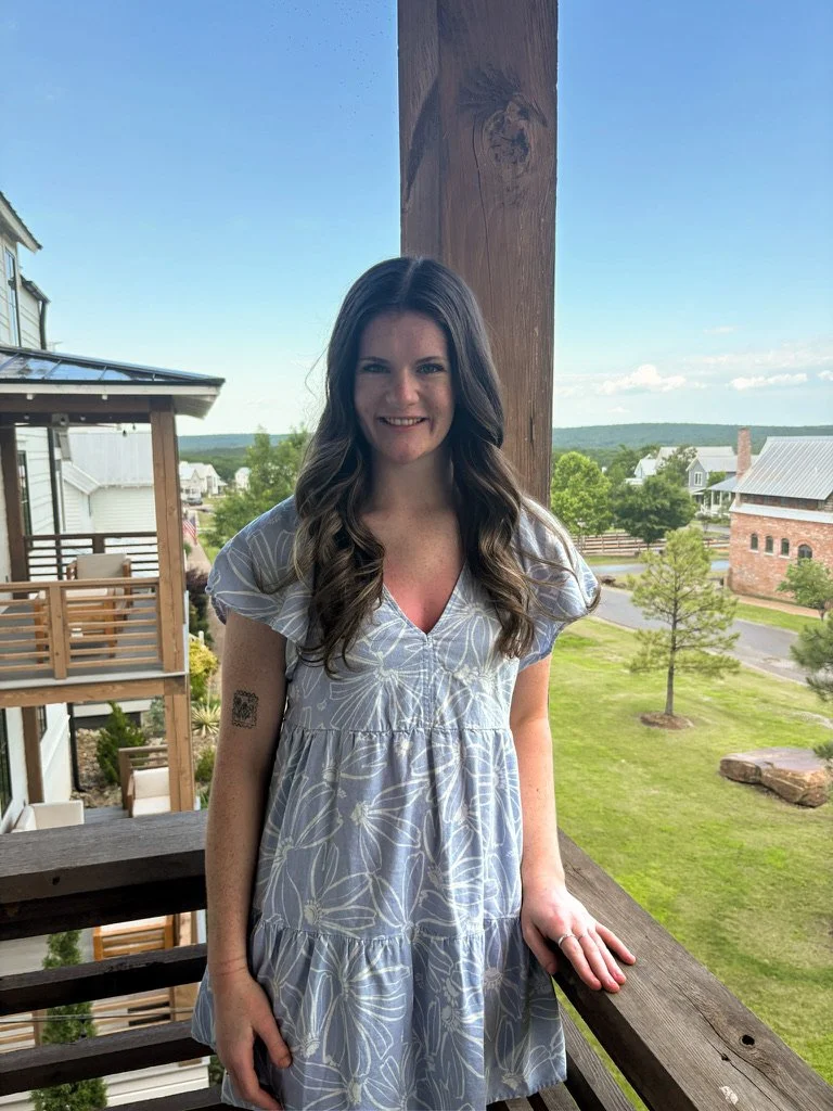 A young woman with long dark hair smiling on a balcony overlooking a grassy area with trees and buildings in the background.