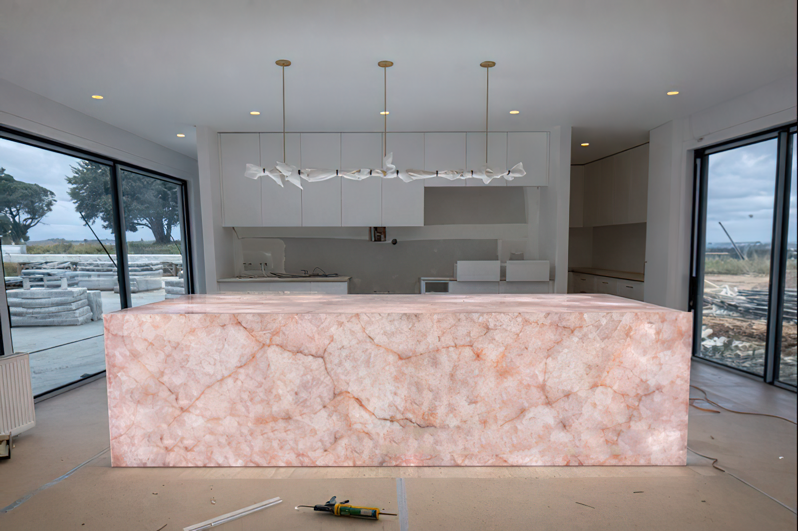 Interior of a modern kitchen under construction with a large pink marble kitchen island, white cabinets, and large sliding glass doors showing outdoor scenery.