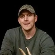 A man wearing a camouflage cap and dark green shirt, smiling with a dark background.