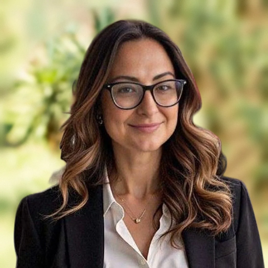 Rae Cee Vallejos smiling, wearing glasses, a black blazer, and a white shirt with green foliage behind her