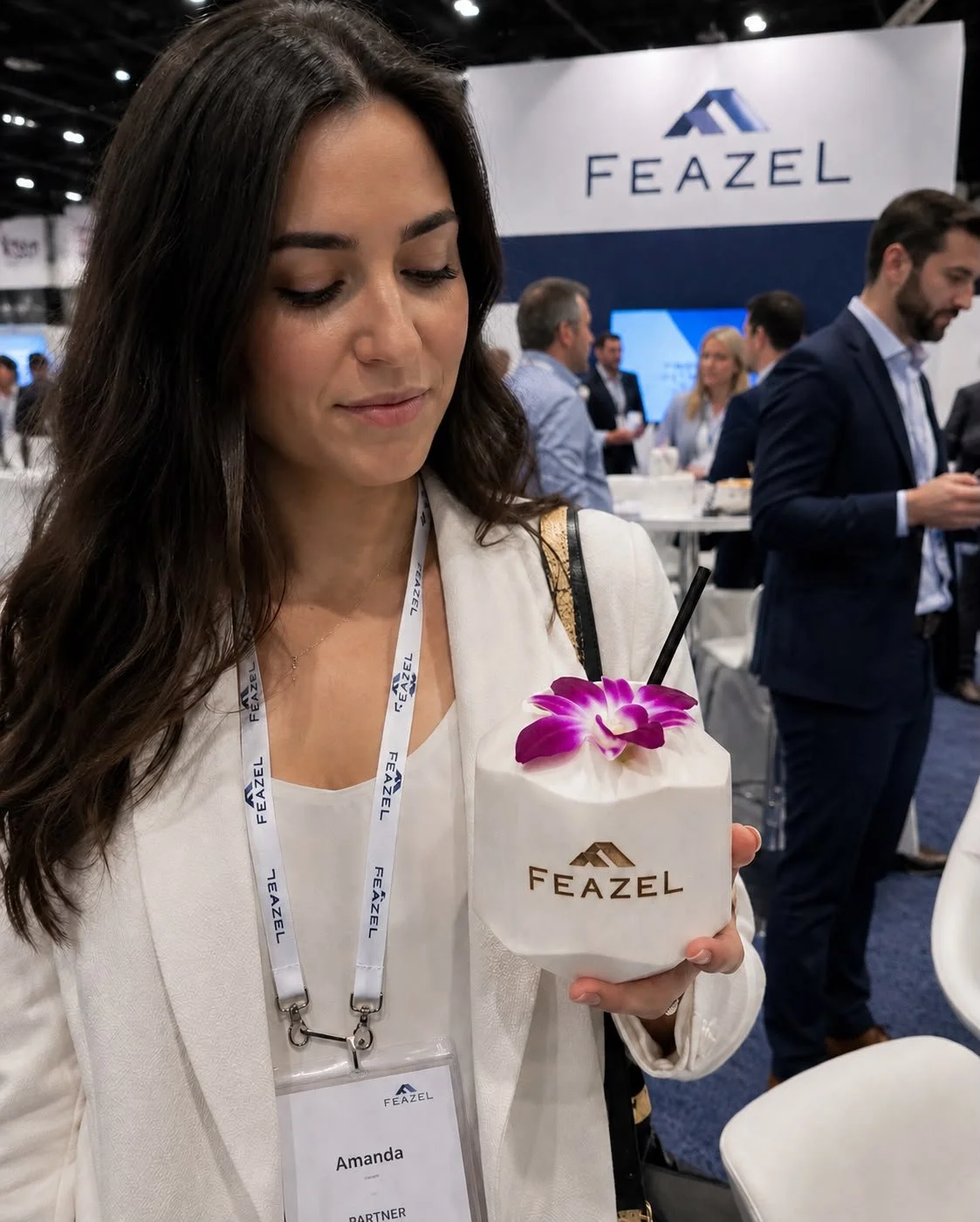 A woman holding a coconut-shaped drink with an orchid flower on top at a trade show with a Feazel sign in the background.