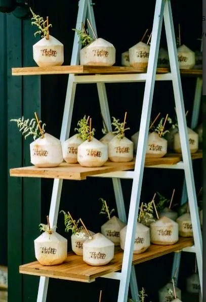 Multiple white coconuts with straw and greenery, displayed on a wooden ladder shelf, with gold writing on the coconuts that says 'Thank you' or 'Thank You'.
