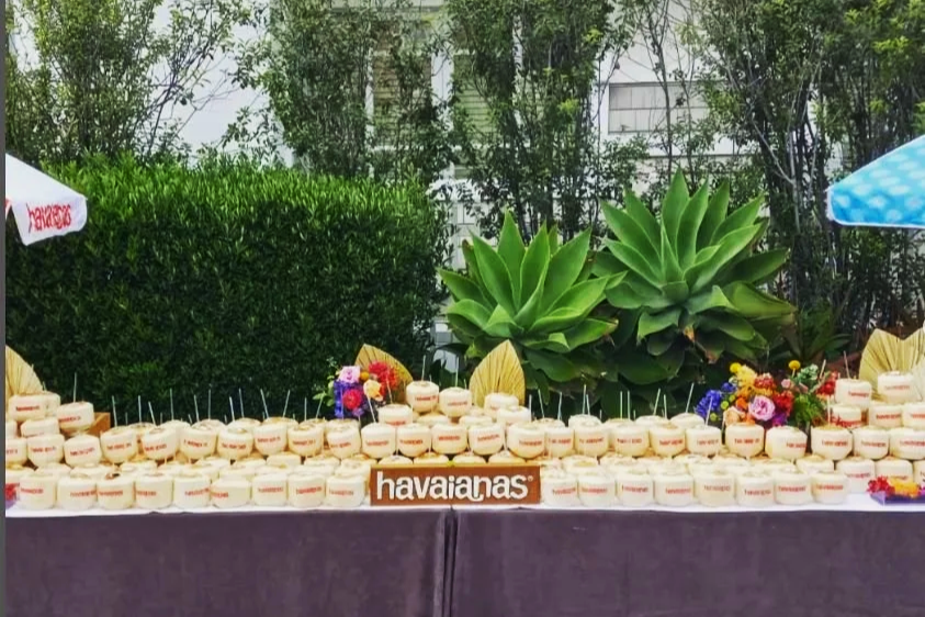 A long table with numerous small cakes, a wooden sign that reads 'havaianas,' and colorful flowers, set outdoors with greenery and large leafy plants in the background.