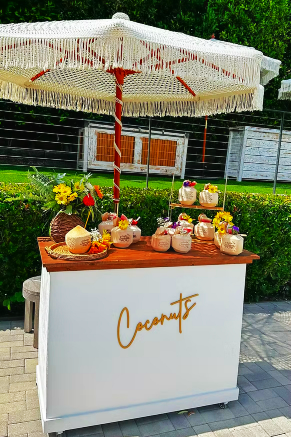 A coconut stand with a large straw umbrella, decorated with tropical flowers, selling coconuts with straws and umbrellas, with a sign reading 'Coconuts' in orange script.