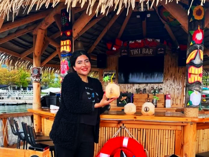 Woman smiling and holding a branded coconut for bars at a tiki bar with tropical decorations and a thatched roof.