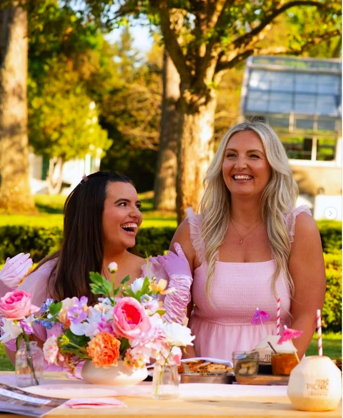Two women in pink dresses laughing and smiling at a birthday party outdoors, with a table of flowers, cake, and custom-branded coconuts for birthday parties, surrounded by trees.