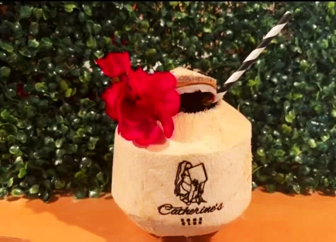 A coconut drink with a striped straw, red flower garnish, and a small umbrella in front of a green leafy background.