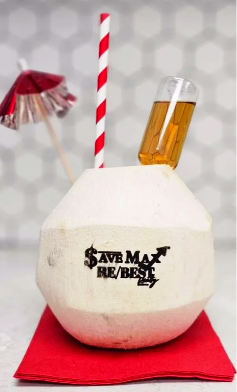 A coconut drink with a straw, small umbrella, and shot glass filled with a yellow liquid, placed on a red napkin.