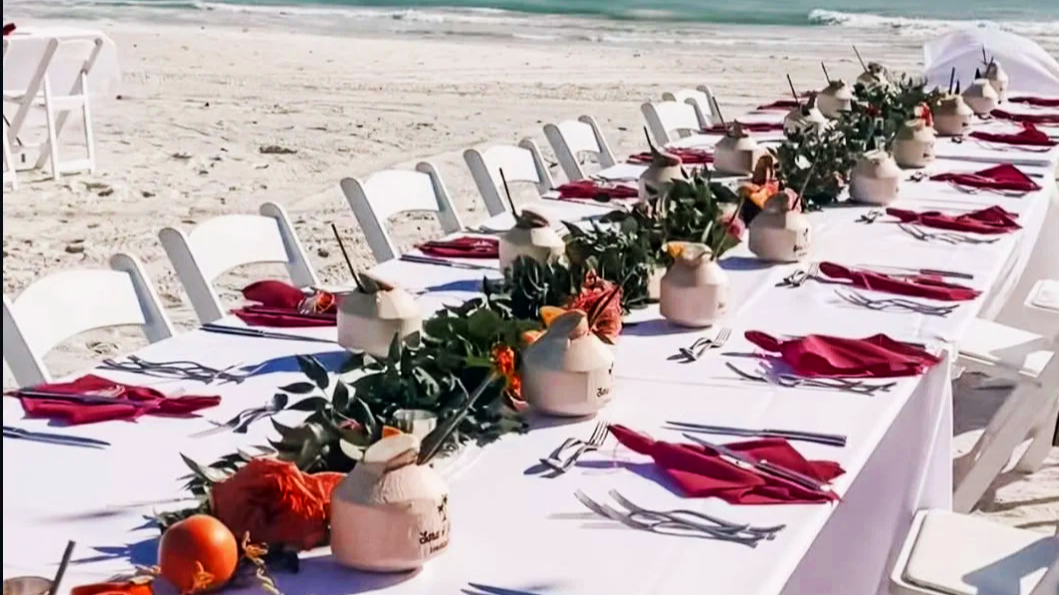 A long beachside dining table set with white tablecloths, red napkins, ceramic jugs, and floral centerpieces with greenery and orange flowers, on a sandy beach near the ocean.
