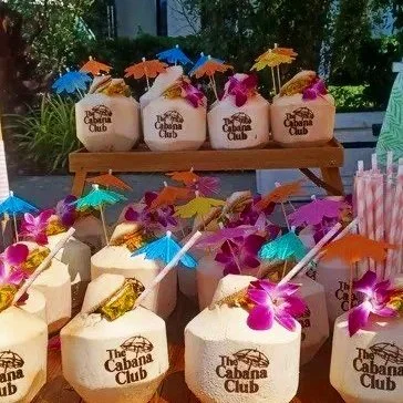 Several coconut drinks with colorful umbrellas and orchids on top, lined up on a table outdoors at The Cabana Club.