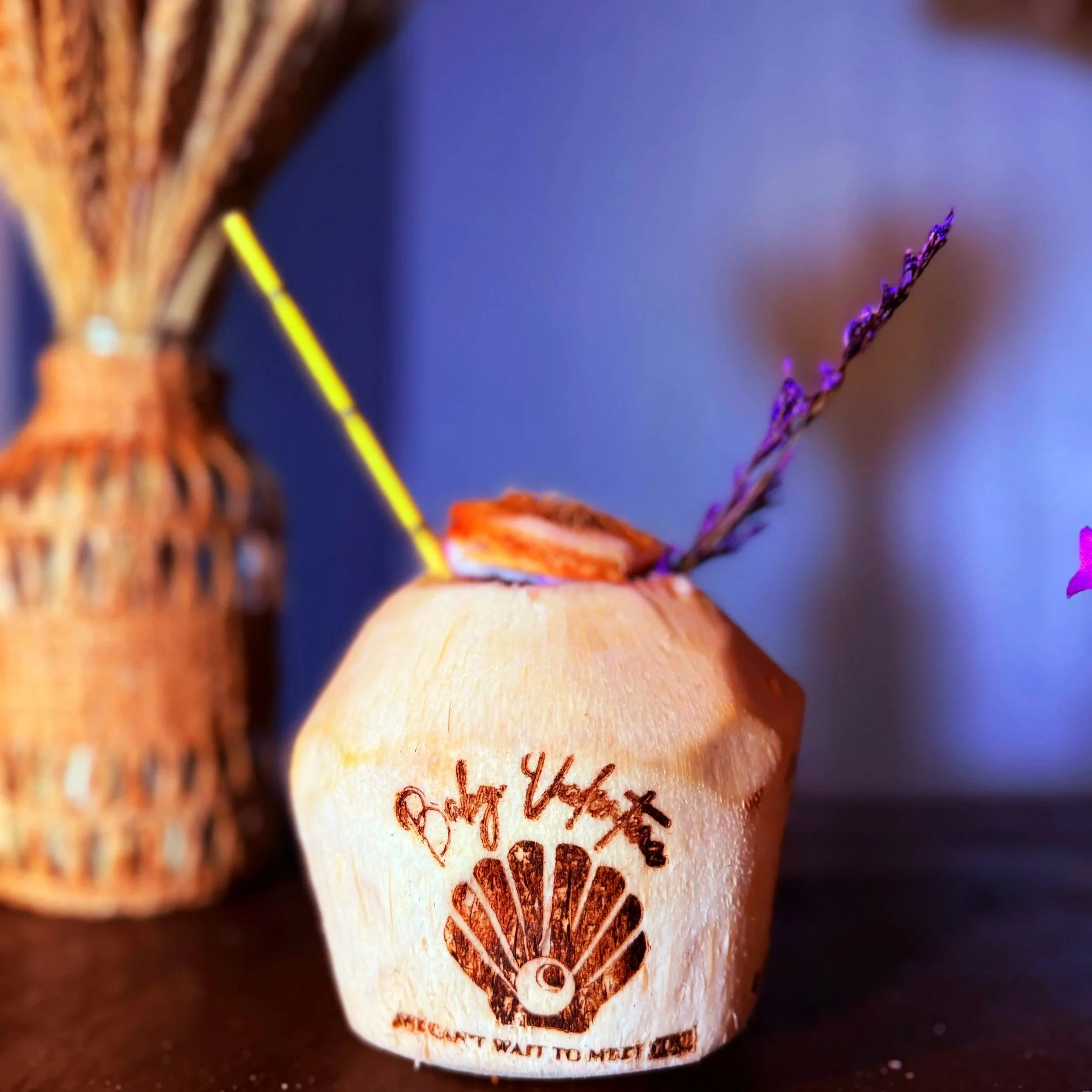 A branded coconut with a logo and text in front of a woven basket in the background. The container has a purple flower and a yellow stick on top.