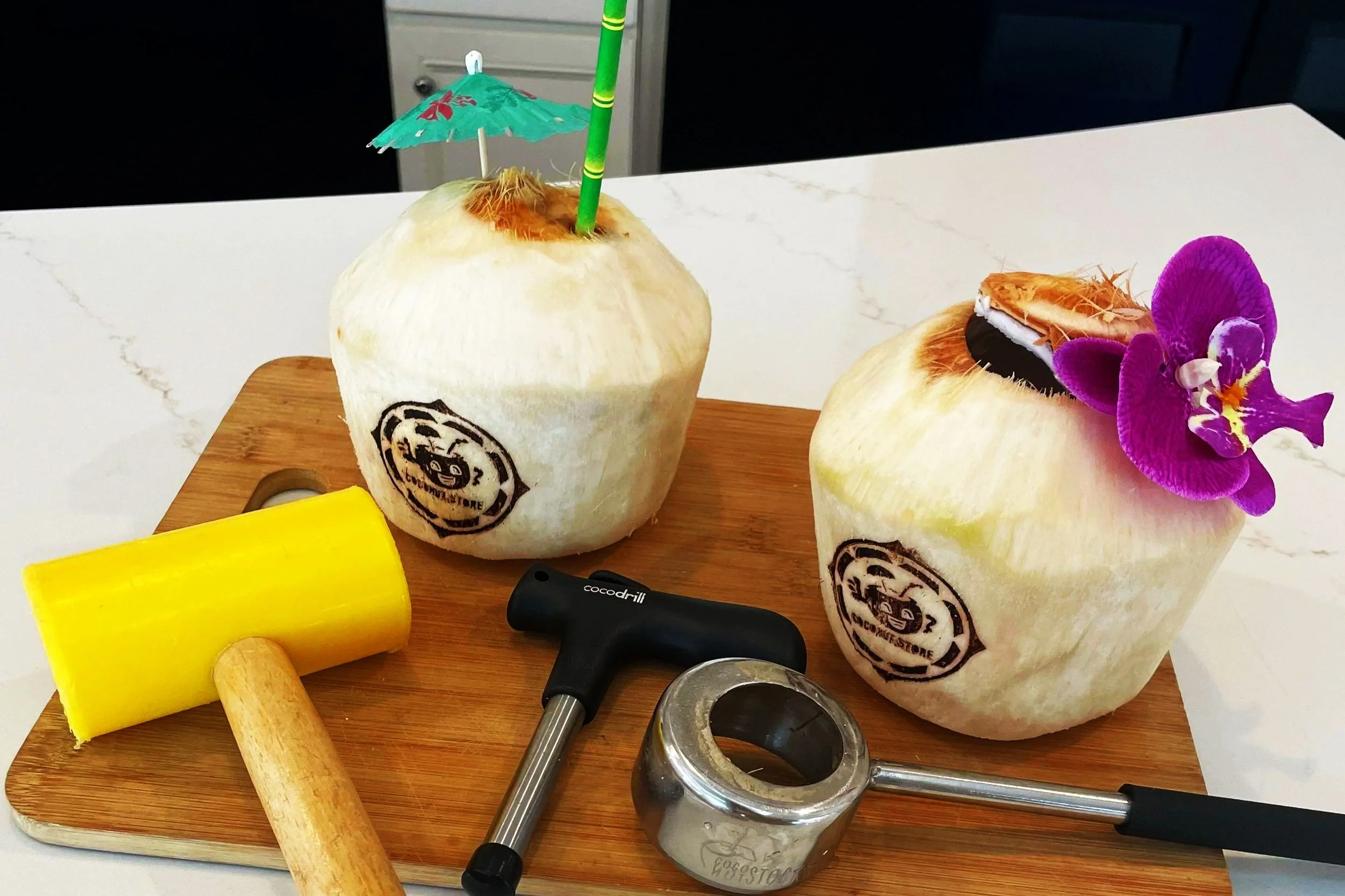 Two young custom-branded coconuts with straws and a coconut opener, placed on a wooden cutting board.