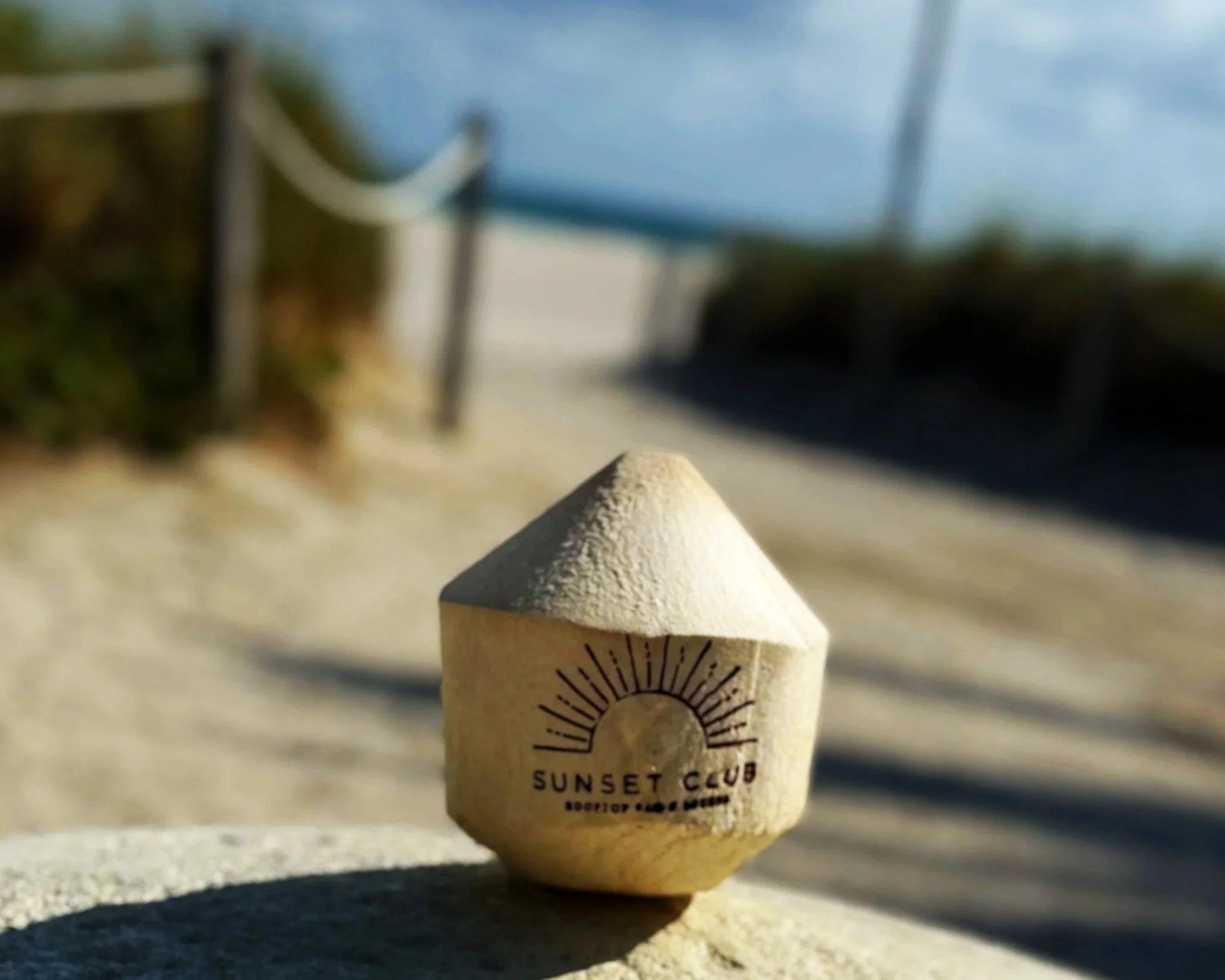 A small beige cone-shaped branded coconuts with a printed sunset, palm trees, and the words 'Sunset Club' on it, placed on a sandy surface at the beach with a blurry background of a fence and sky.
