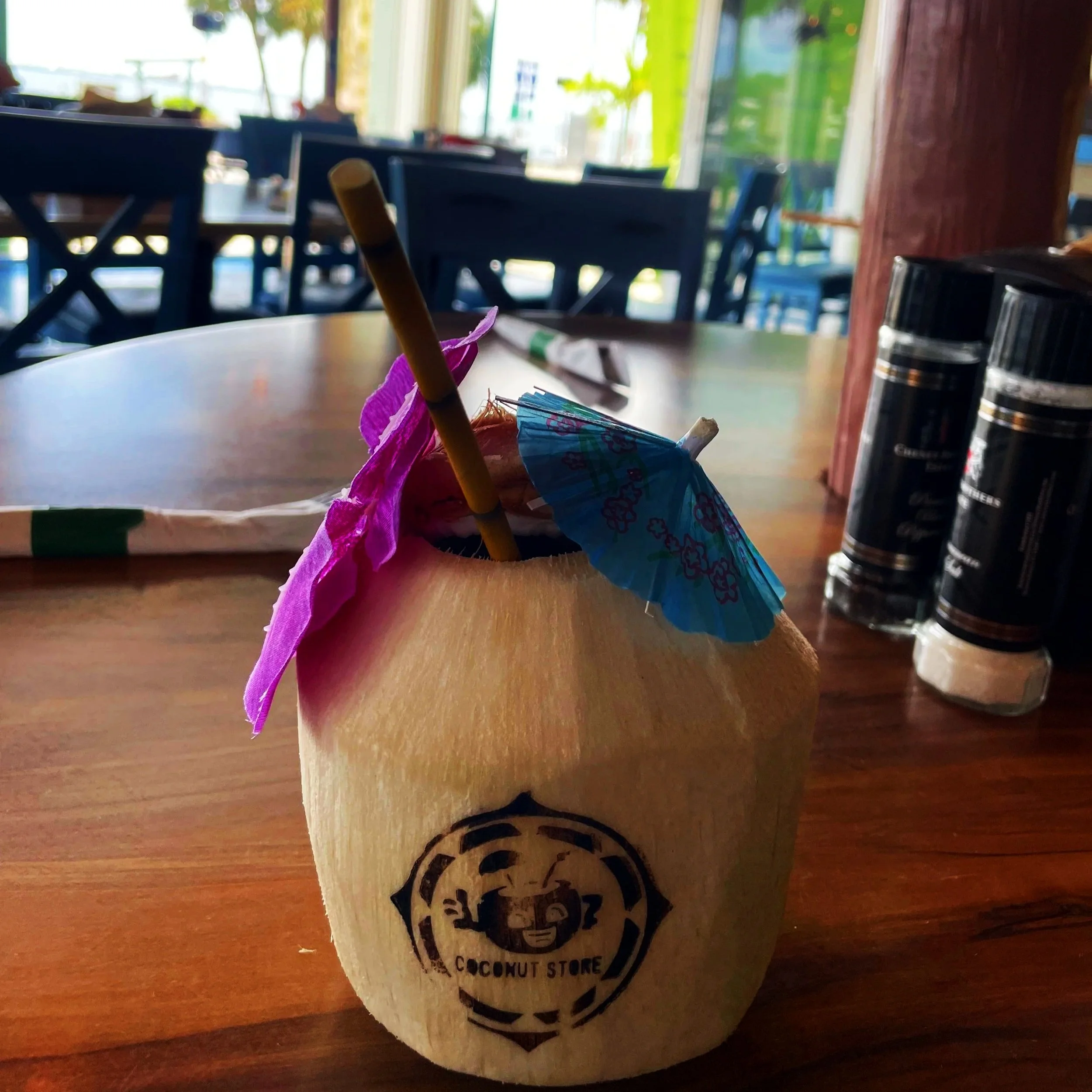 A fresh coconut drink decorated with pink and purple paper umbrellas and a blue cocktail umbrella, placed on a wooden table inside a restaurant or cafe.
