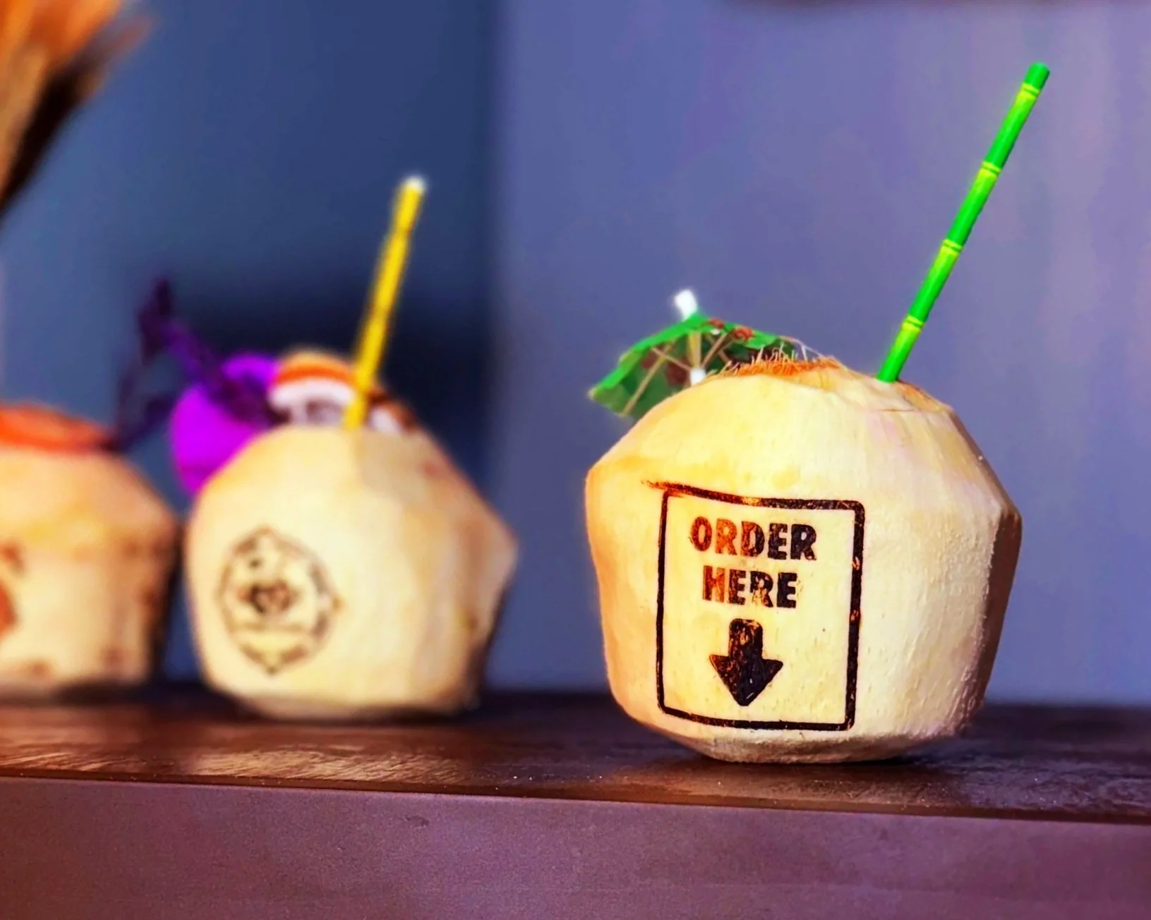 Miniature coconut drinks with colorful umbrellas and straws on a wooden surface. One has a label that says 'Order Here' with a downward arrow.