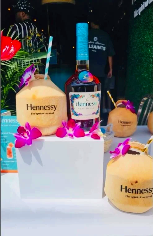 Various bottles of Hennessy cognac with coconut-shaped labels and pink orchids on a table, along with coconut drinks with striped straws, at a tropical-themed event.