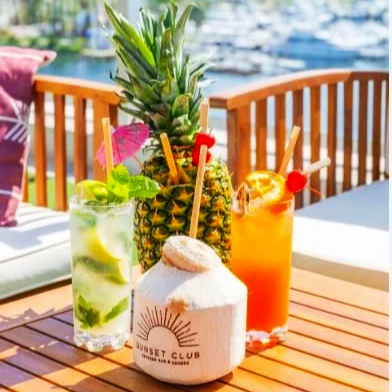 Four tropical drinks with garnishes and a pineapple on a wooden table outdoors by the water.