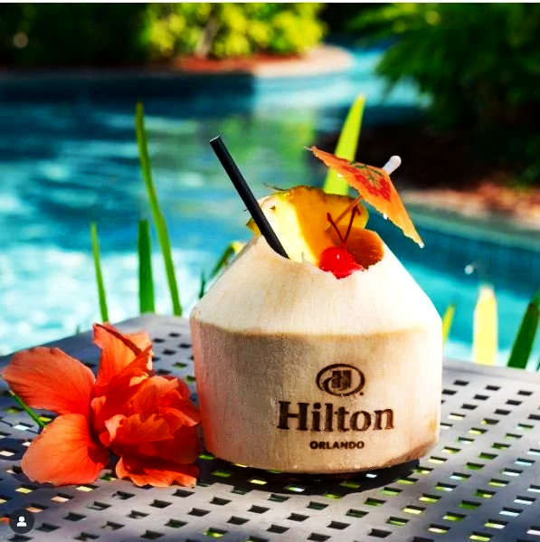 A branded coconut drink with an umbrella, cherry, and straw on a table near a pool, with orange flowers in the foreground and tropical greenery in the background.