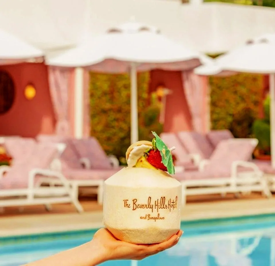 A hand holding a coconut with a labeled logo that reads 'The Beverly Hills Hotel and Bungalows', with pink lounge chairs, umbrellas, and trees in the background.