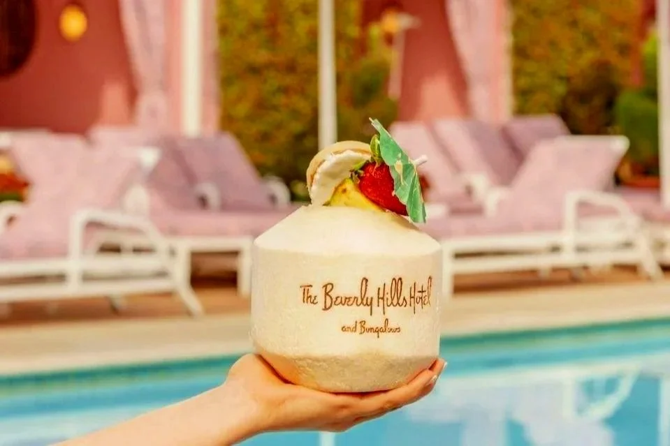 A hand holding a fresh Instagrammable drink with a straw, straw hat, strawberry, and decorative umbrella, with a pool and poolside lounge chairs in the background.