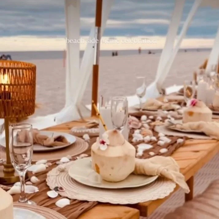 Coastal beachside dining table with shells, candles, and drinks, set under a large white canopy near the ocean.