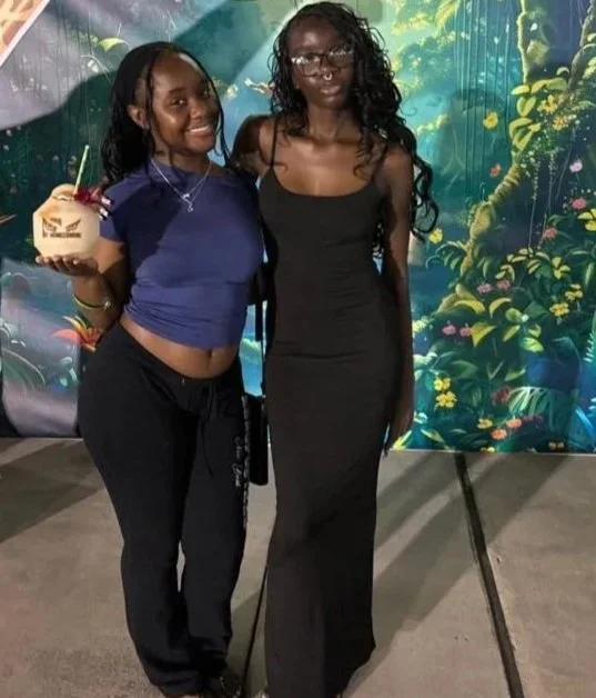 Two women standing in front of a jungle-themed backdrop. The woman on the left is holding a coconut drink.